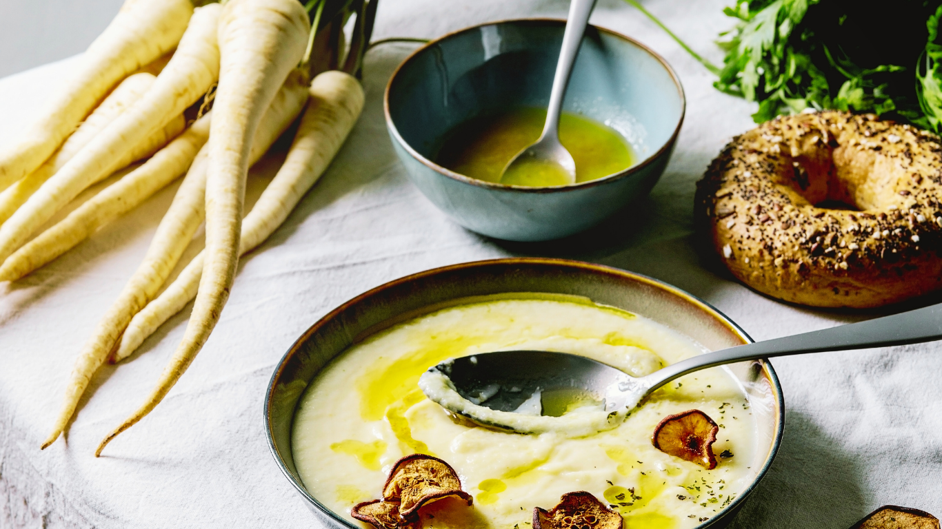 “Creamy parsnip and celery root bisque served in a bowl, drizzled with olive oil and topped with parsnip chips, with a spoon ready to serve.”